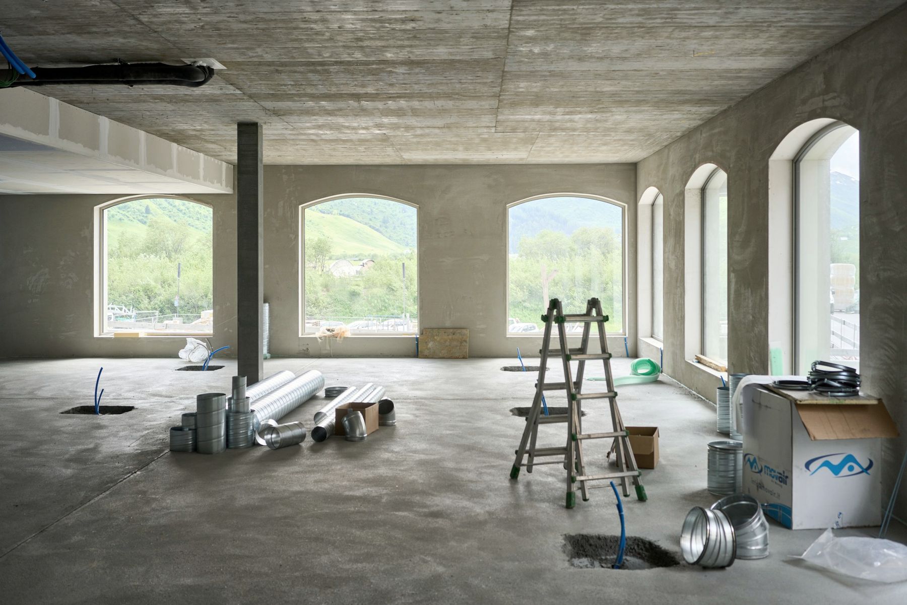 An unfinished concrete floor with arched windows, a stepladder and ductwork ready to fit